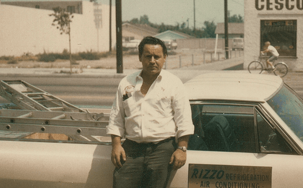 Camille Rizzo, who founded the company in 1963, in a white work shirt leaning against a Rizzo Refrigeration & Air Conditioning service car parked on a sunlit street beside a CESCO storefront
