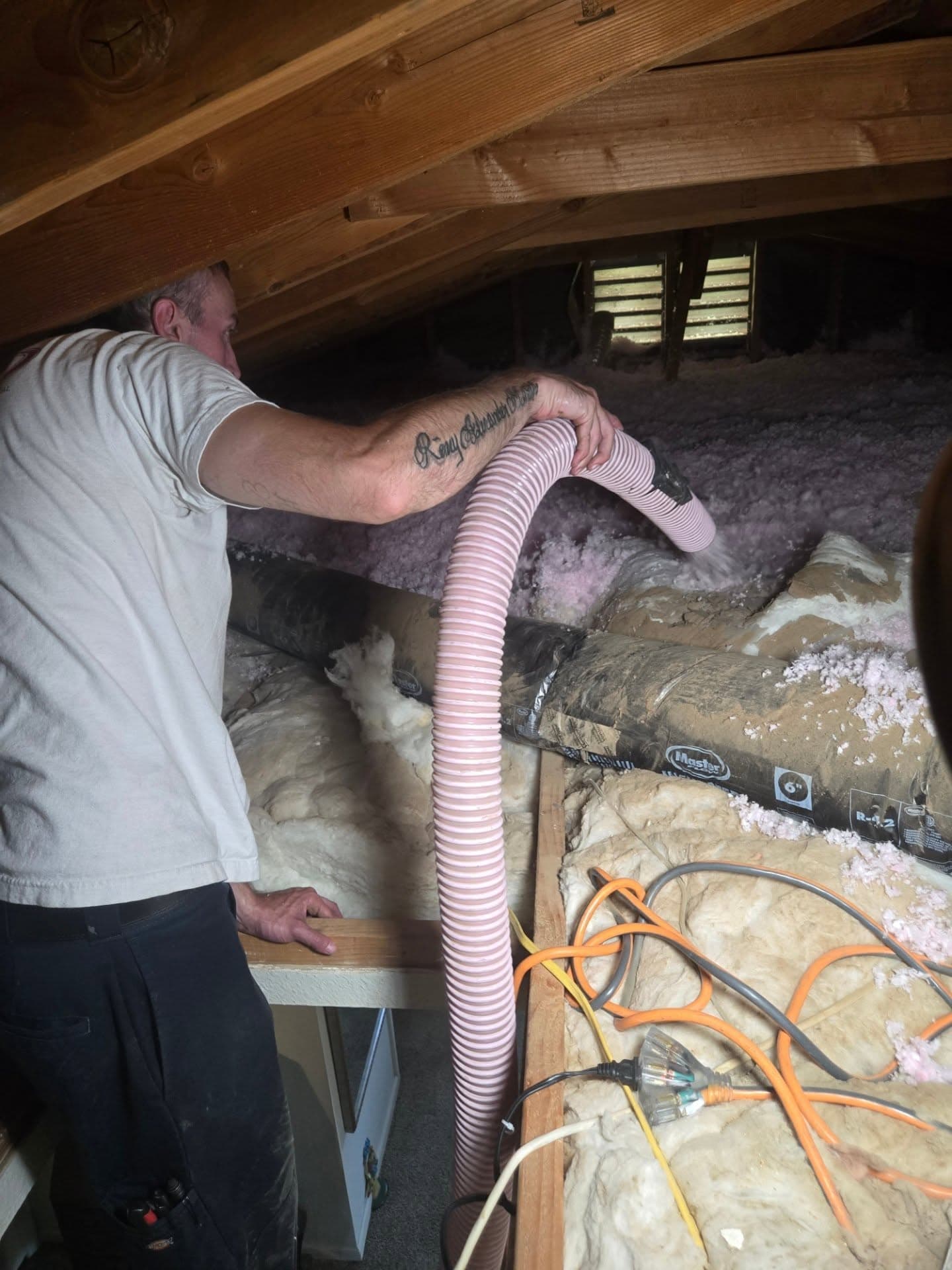 A Rizzo technician kneeling at an attic hatch, feeding a pink blower hose over the joists and laying loose-fill insulation across the old batts and duct runs