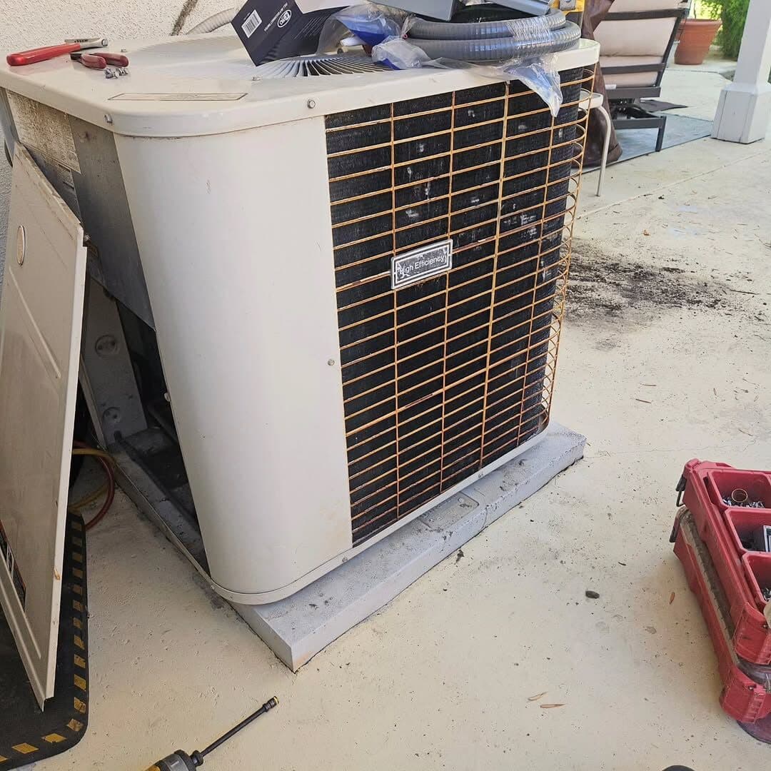 The old condenser on its pad, coil fins corroded orange, panel off and tools on the patio