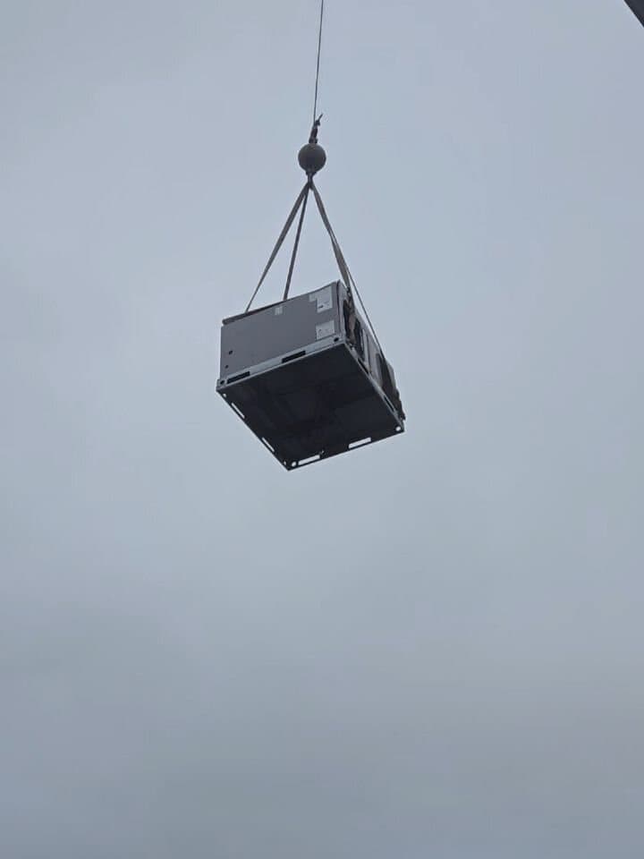 A package unit hanging from a crane hook against an overcast sky